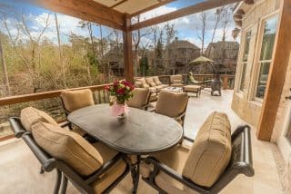A spacious patio has beige cushioned chairs, a black table with flowers, cozy seating, trees behind, and nearby houses beyond a fence.