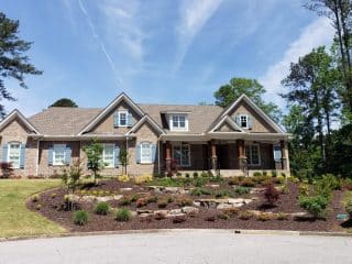 Planting beds with shrubs, rocks and open soil extend across the front of a house beneath a bright blue sky.