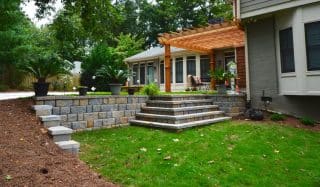 A backyard with a stone retaining wall, wide steps, pergola-covered patio, potted plants, green grass, and a house with large windows and trees.