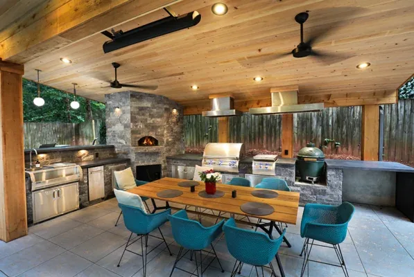 An outdoor kitchen with a wooden ceiling, blue dining chairs, grills, pizza oven, stone accents, and lighting on a tiled patio in a fenced yard.