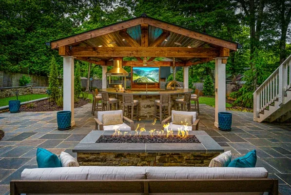 A cozy stone patio has cushioned seats, a fire pit, and a covered bar with stools and a TV, with trees and greenery around it.
